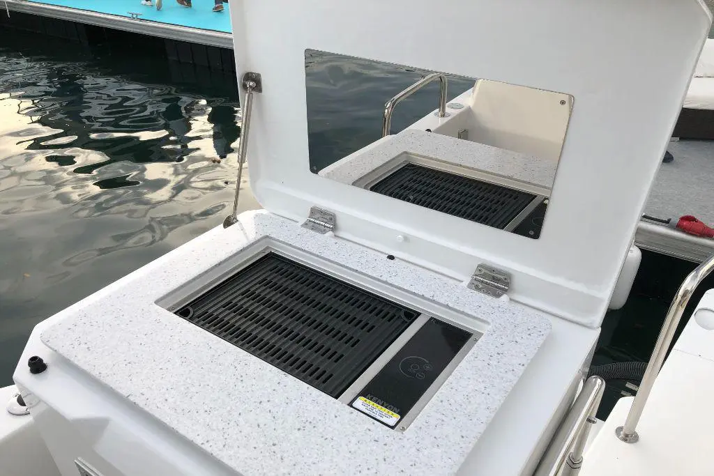 Boat BBQ grill with a white surface, open lid showing a black grill. Calm water and a pontoon are visible in the background.